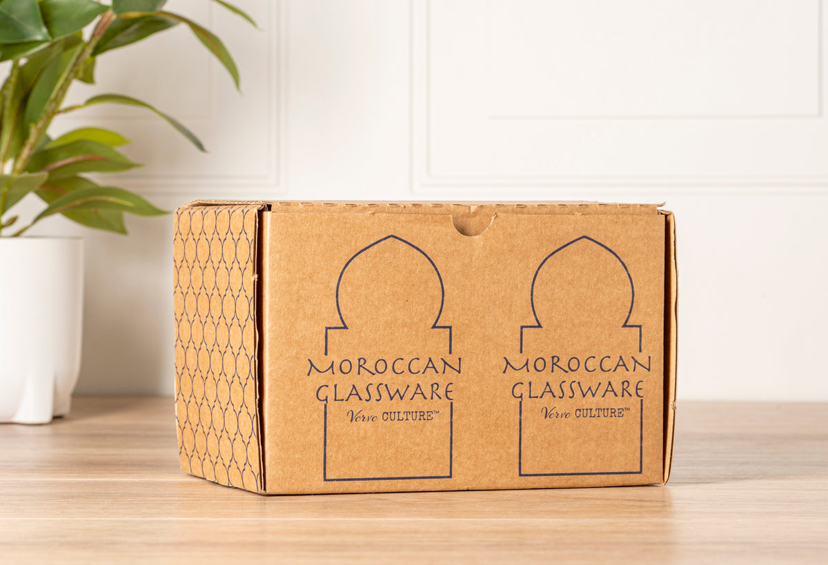 Cardboard box with geometric pattern containing Moroccan cone-shaped glassware set on wooden surface next to potted plant.