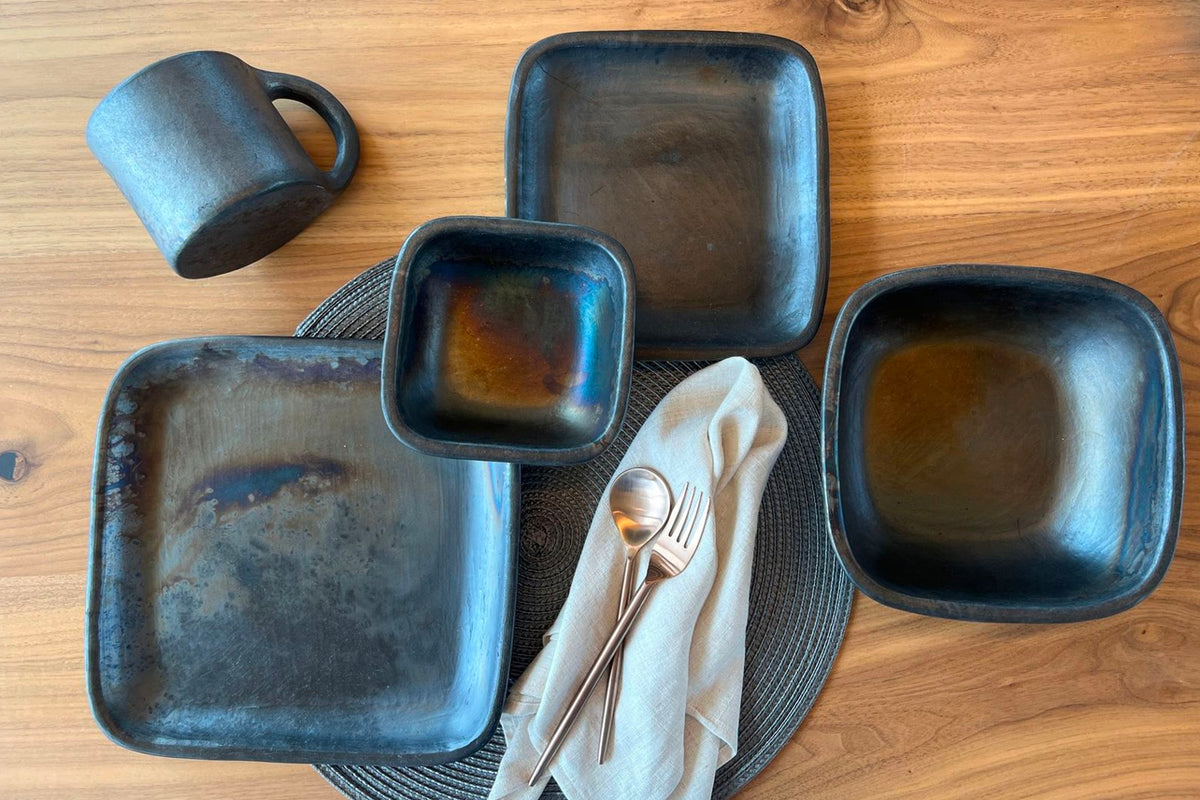 Dark blue-grey handmade pottery square bowls in varying sizes arranged on a wooden surface with a mug and utensils.