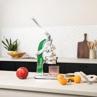 Green and silver citrus juicer with lever arm, positioned on countertop next to red apple, small oranges, and glass collectio