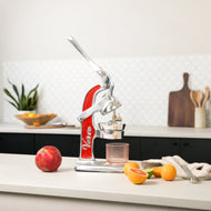 Red and silver manual citrus juicer with lever handle, positioned on kitchen counter next to whole and halved citrus fruits.