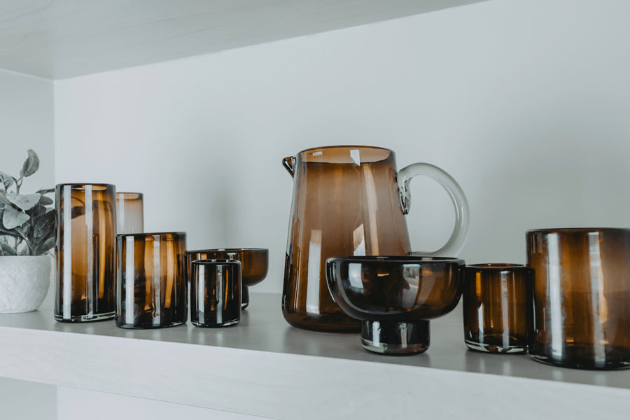 Clear and amber-toned glass pitcher with handle displayed among matching glassware on white shelf.