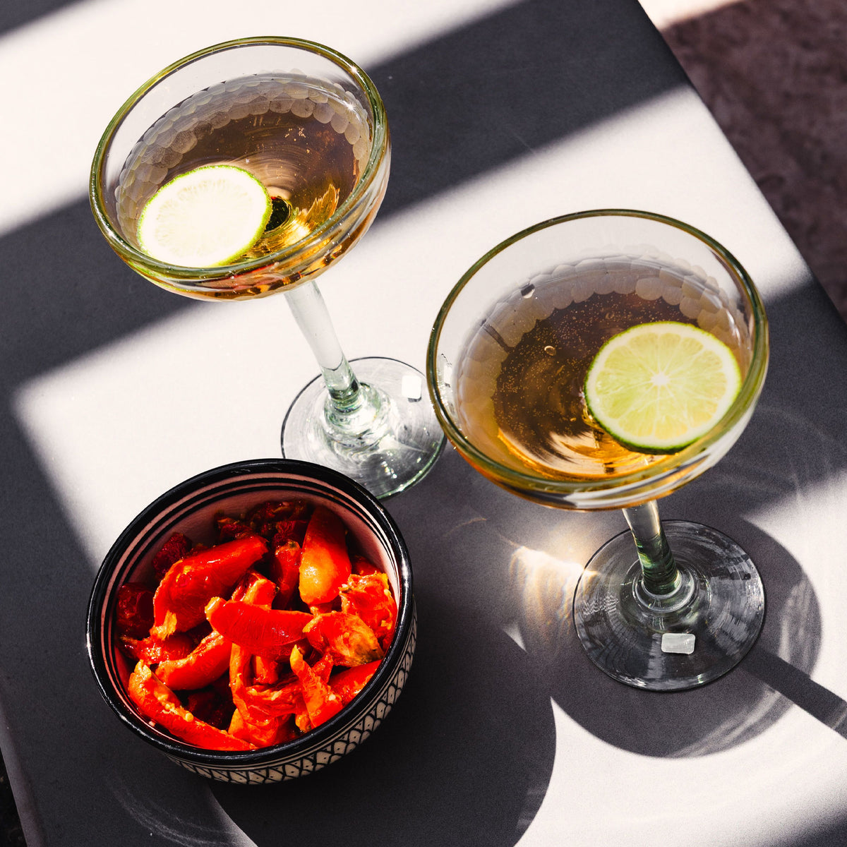 Two clear cocktail glasses with long stems and textured bowl design, one containing amber liquid with lime slice, the other w