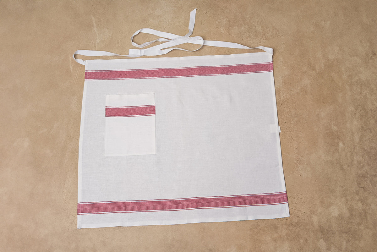 White linen apron with red stripes at top and bottom, front pocket with red trim, and white neck and waist ties.