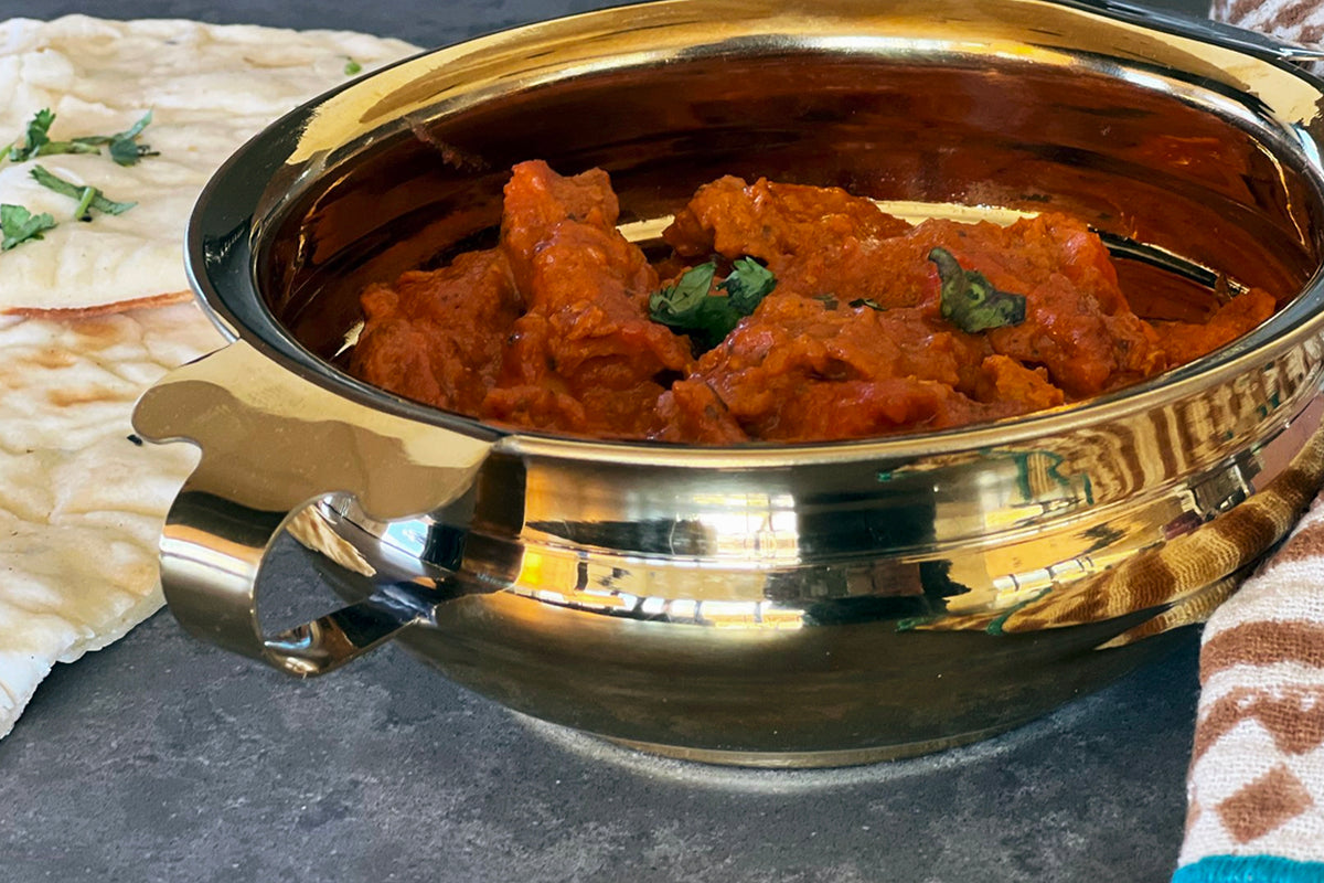 Brass-colored stainless steel handi bowl filled with red curry and meat garnished with cilantro, with handles on sides.