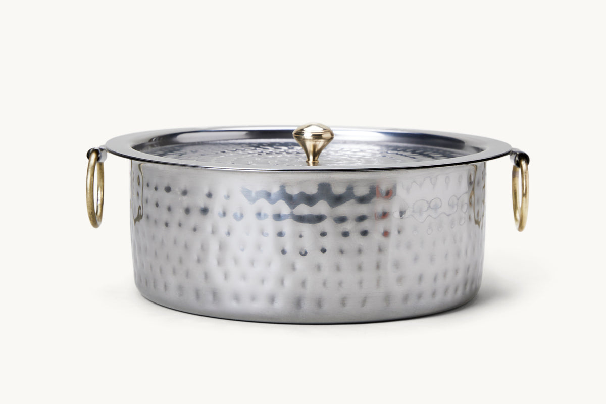 Stainless steel patila pot with hammered surface texture, brass loop handles, and matching lid with brass finial.