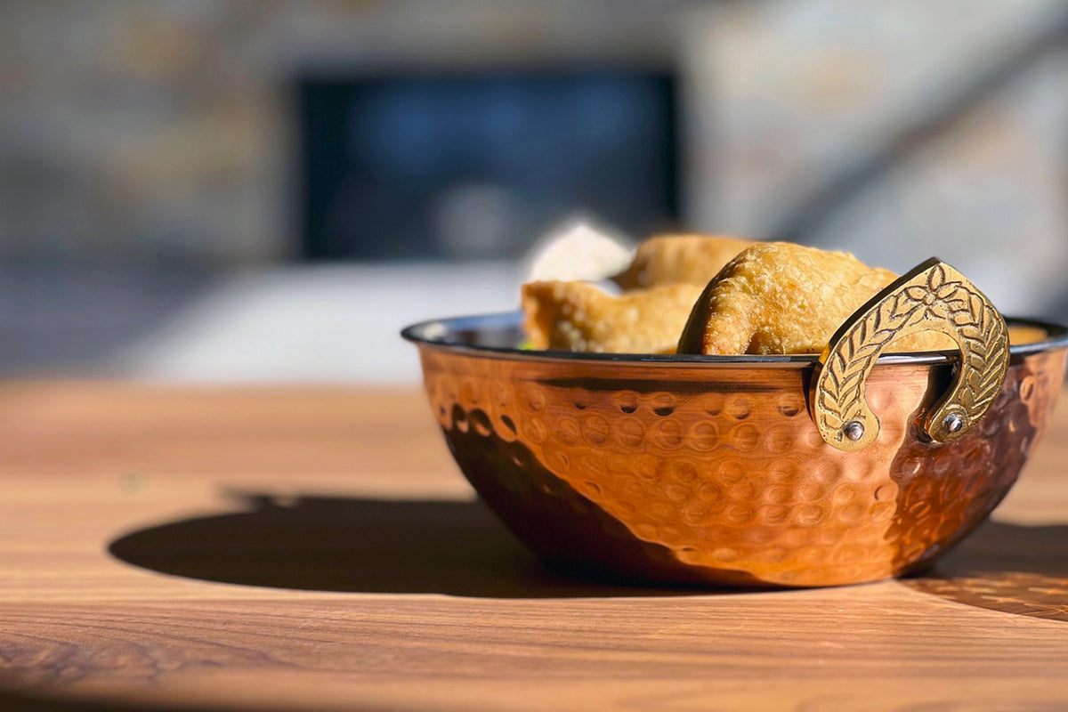 Hammered copper bowl with ornamental handles containing bread, displayed on wooden surface.