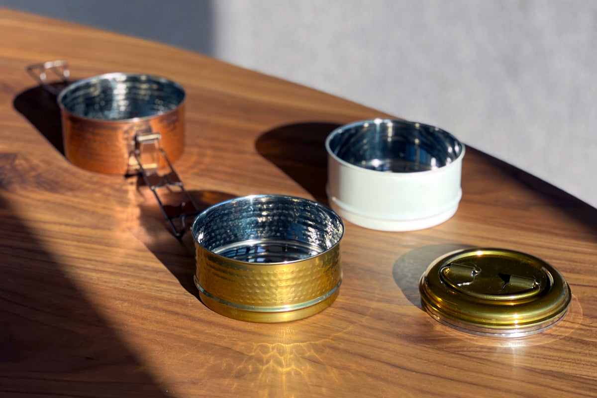 Three stainless steel containers with copper, white, and gold finishes and a brass lid displayed on a wooden surface.