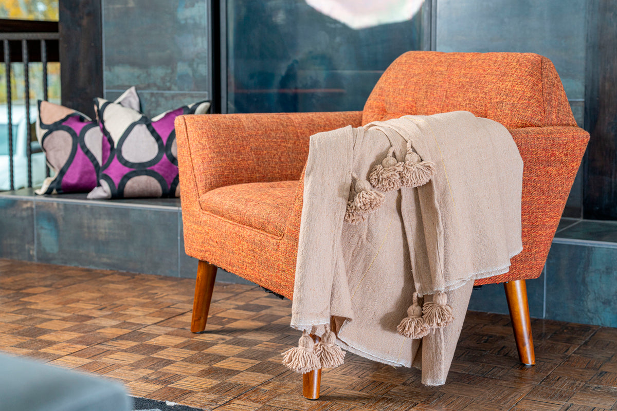 Cream-colored tasseled blanket draped over an orange upholstered chair with wooden legs.
