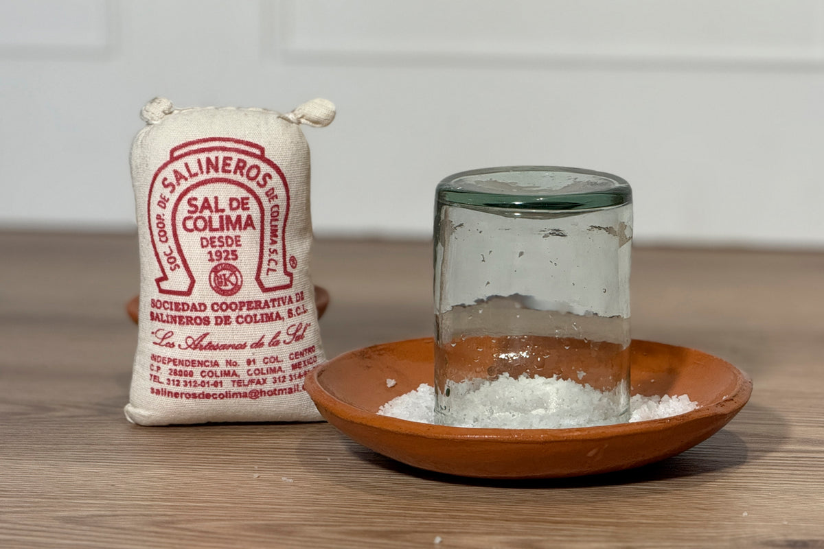 Cloth bag of Colima sea salt with red label next to glass jar and wooden dish containing white salt crystals.