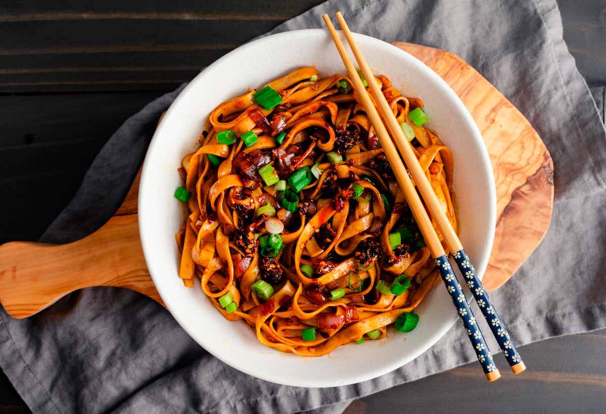 Sweet potato noodles topped with chili crisp, green onions, and red pepper in white bowl with chopsticks.