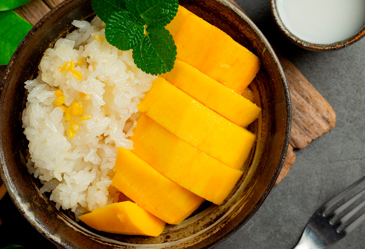 Sticky rice with mango slices and mint leaf in a wooden bowl with coconut milk on the side.