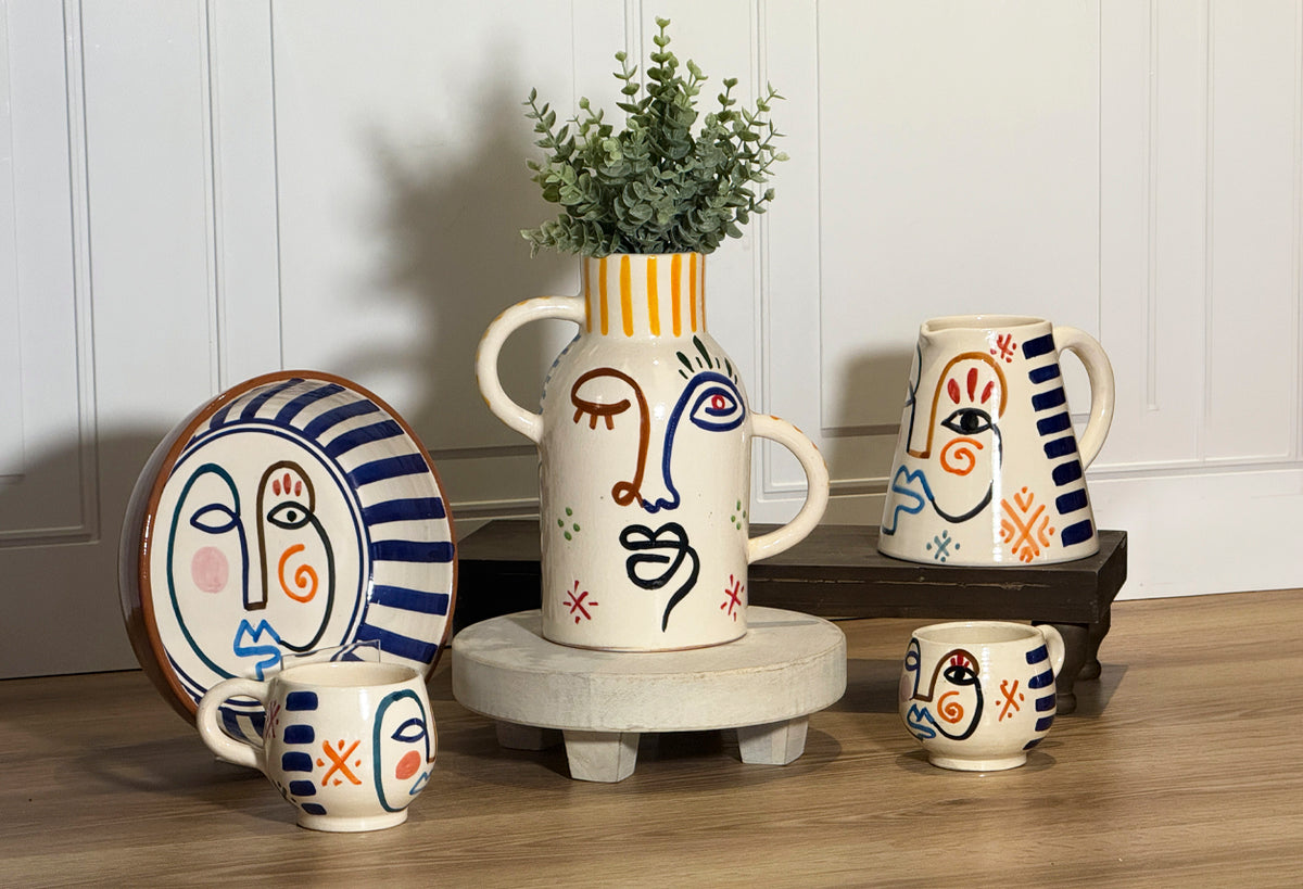 Cream ceramic pitcher with hand-painted abstract face design in blue, orange and red, displayed with matching plates and vess