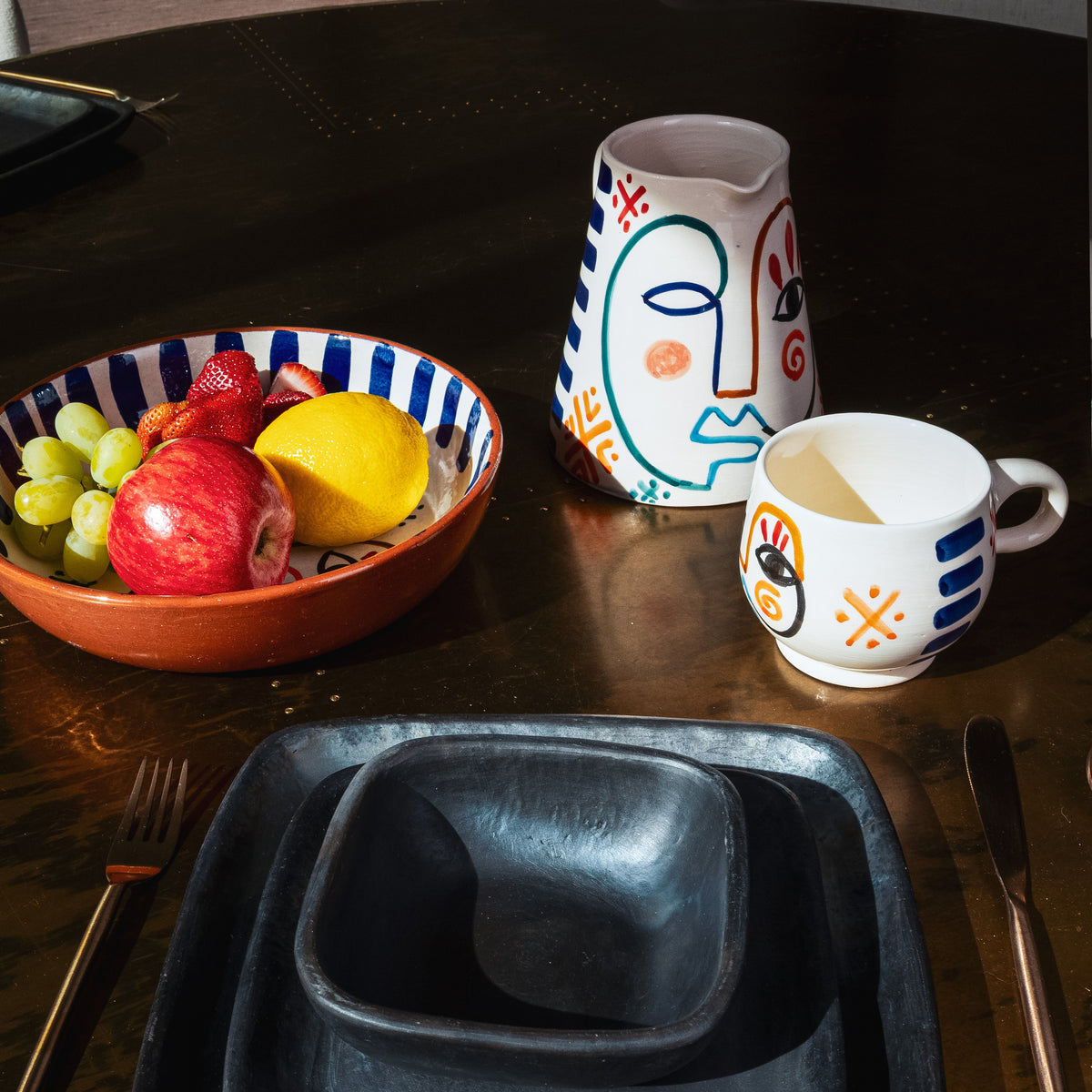 Terracotta bowl with blue and red striped pattern containing fresh fruit, displayed on dark table with painted ceramic vessel
