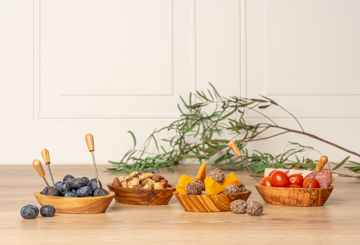 Six small forks with olive wood handles displayed in wooden bowls filled with appetizers and fresh herbs.
