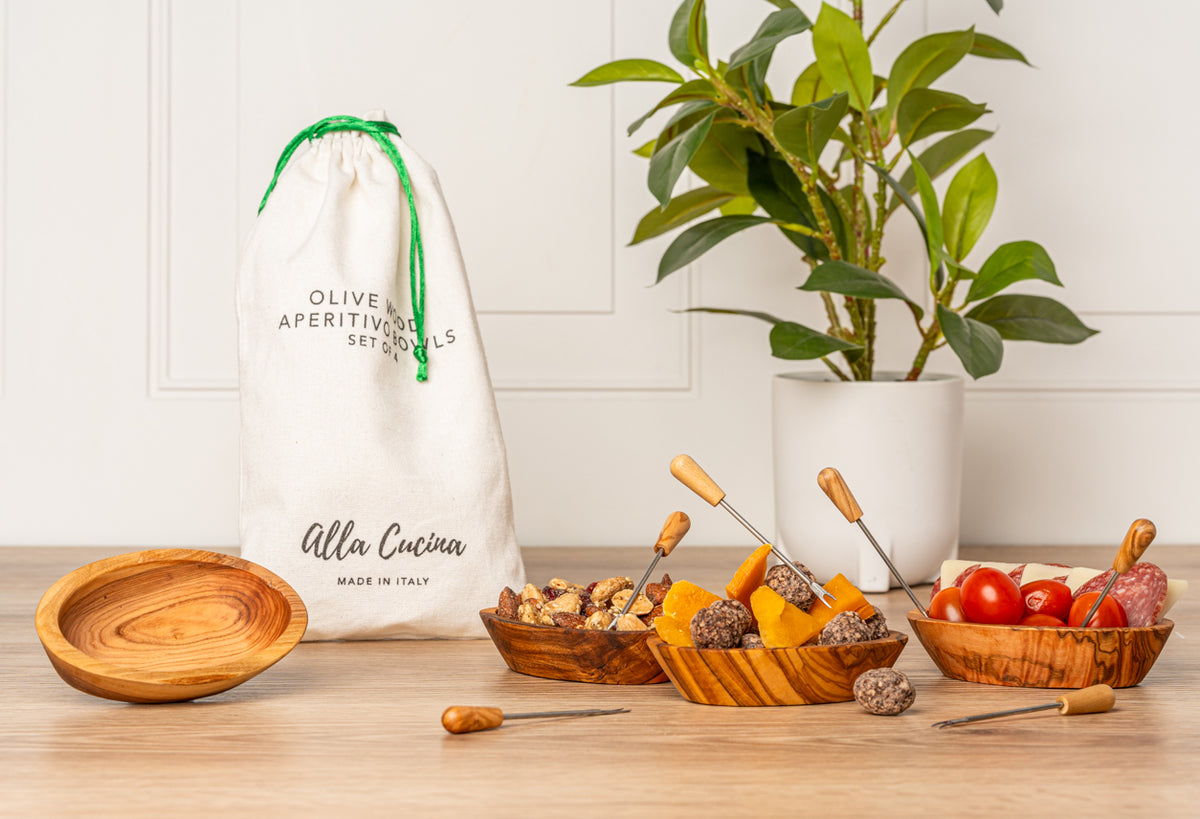 Four wooden bowls with metal serving utensils displayed with olives, cheese, and tomatoes next to a drawstring storage bag.