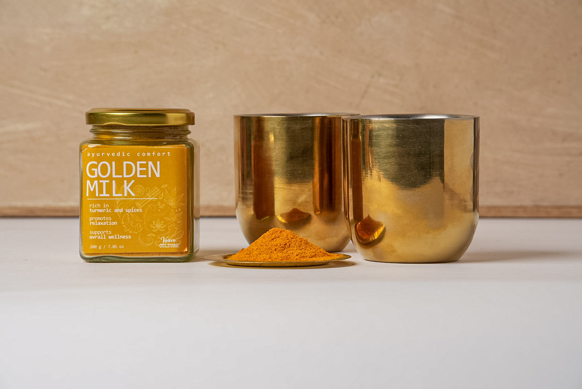 Golden milk spice blend powder in glass jar with gold lid, displayed with two gold metallic cups and a pile of turmeric-color