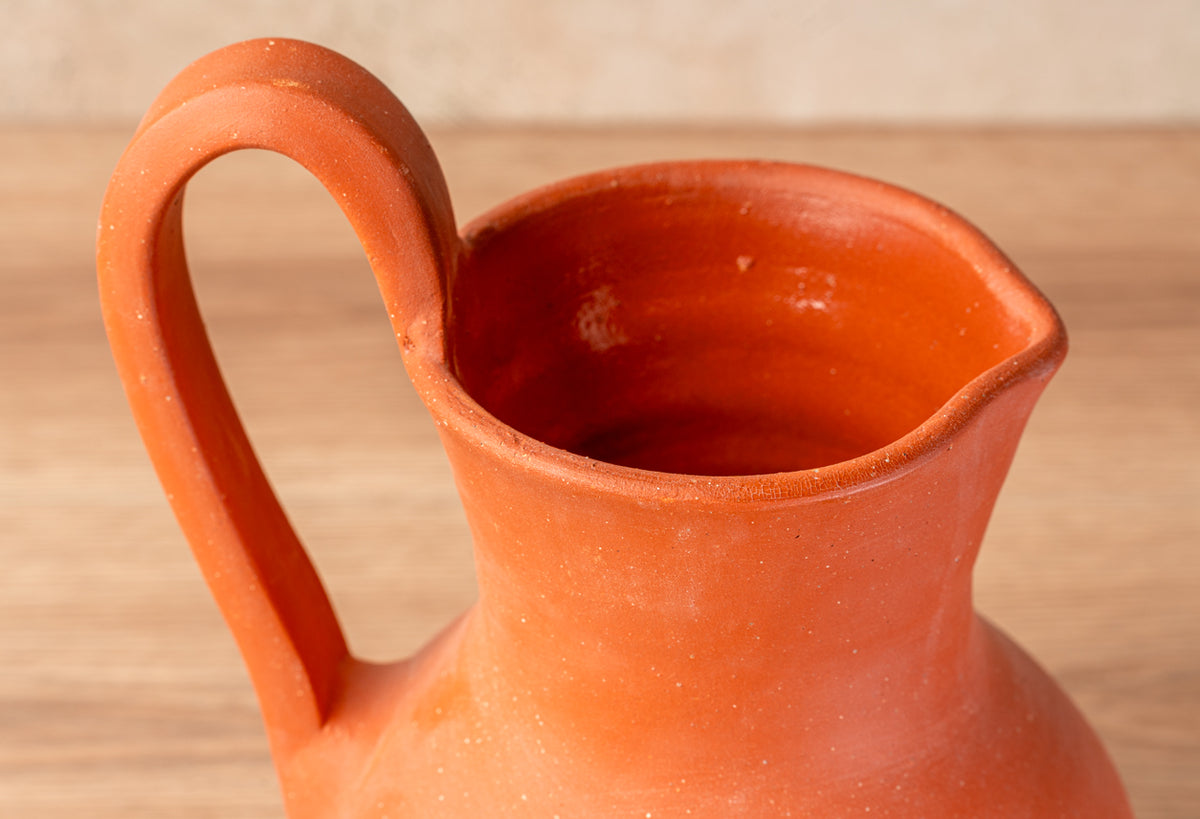 Orange ceramic jug with curved handle and pouring spout on wooden surface.