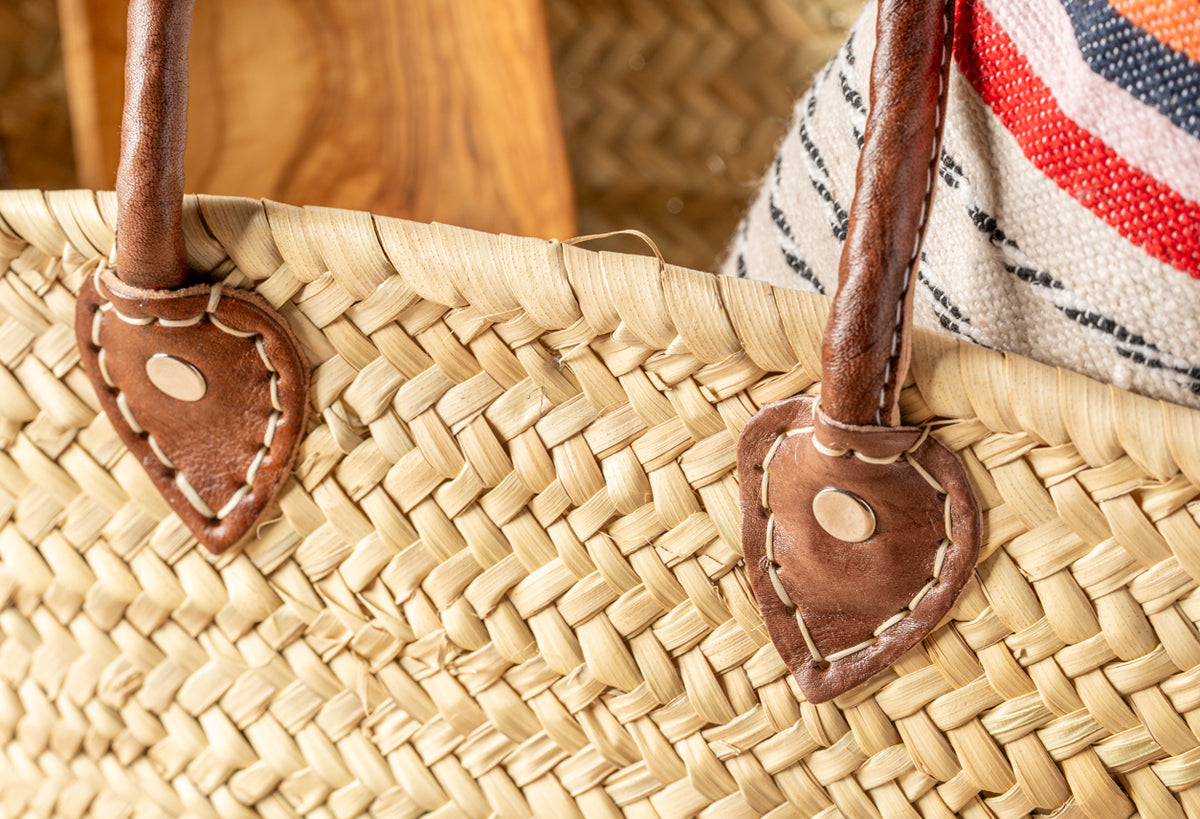 Woven straw basket with brown leather handles and striped fabric trim in red, white, and navy.