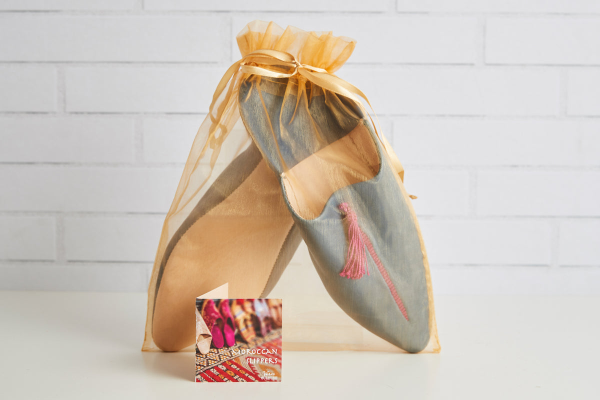 Pair of leather slippers with pointed curved toes, tan and gray coloring, pink tassel detail, displayed in yellow organza bag