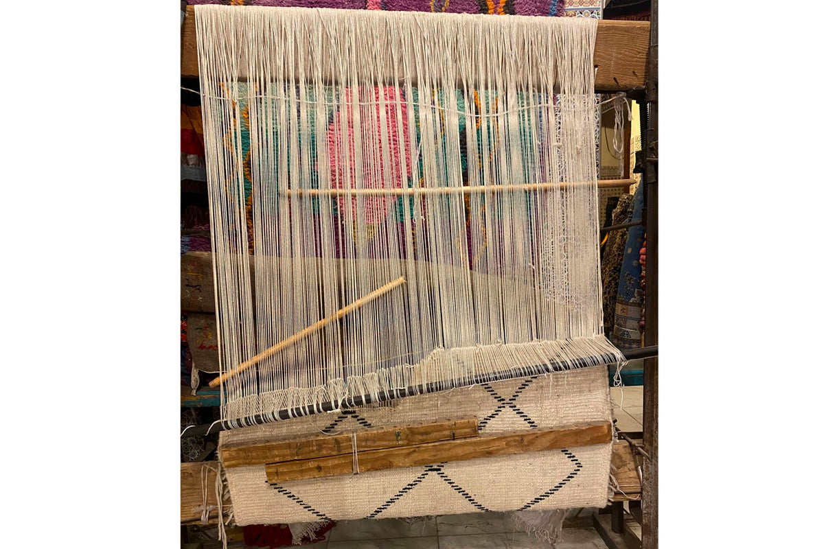 Handwoven rug on wooden loom showing cream base with geometric diamond pattern and colorful vertical stripes in progress.