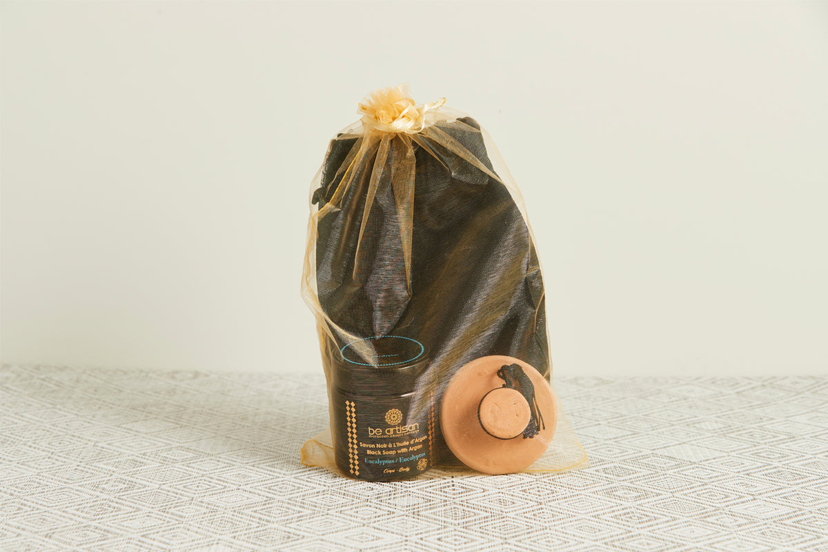 Black drawstring pouch with gold ribbon tie containing dark jar and terracotta bowl with lid on woven surface.