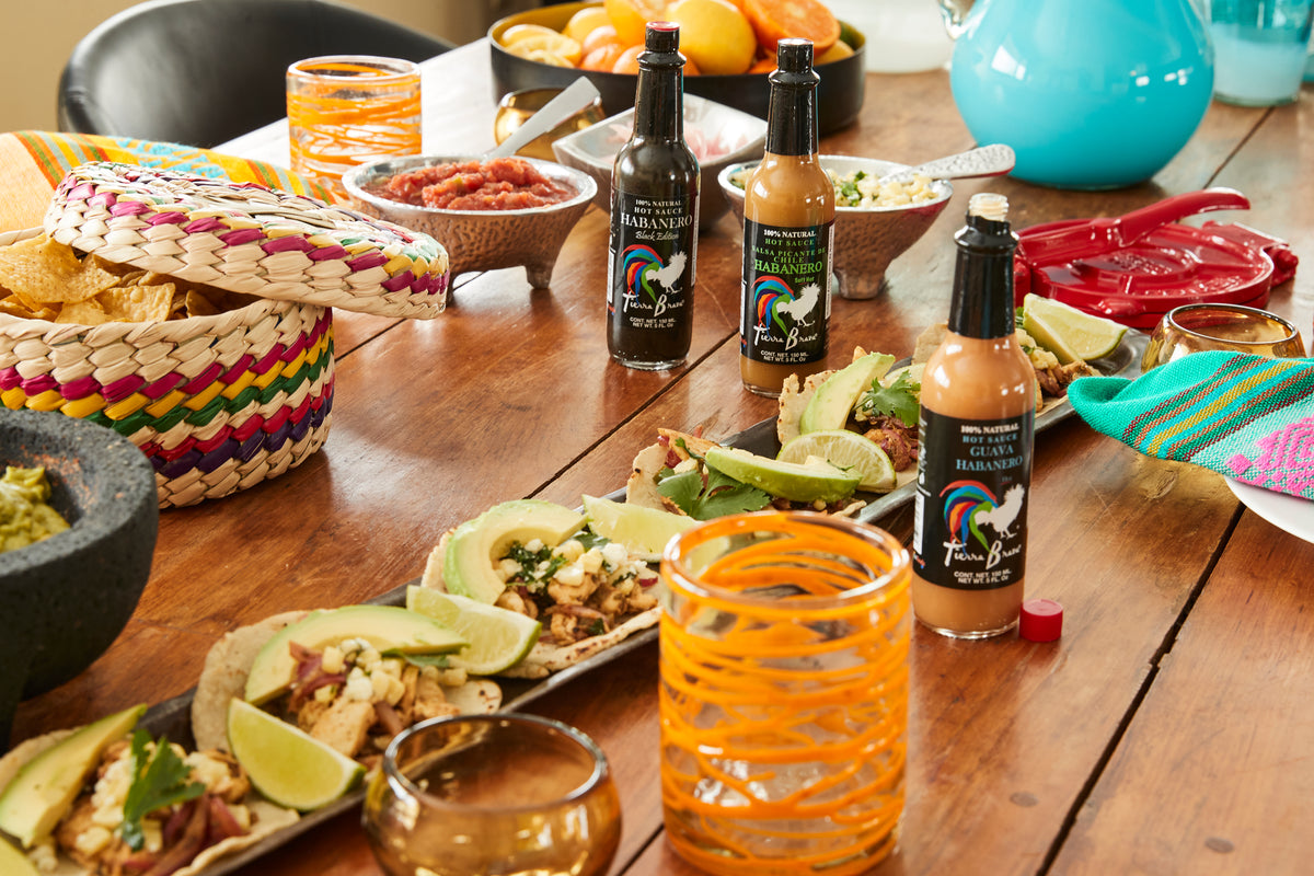 Taco ingredients and condiments displayed on wooden table with woven baskets, hot sauce bottles, fresh limes, and colorful se