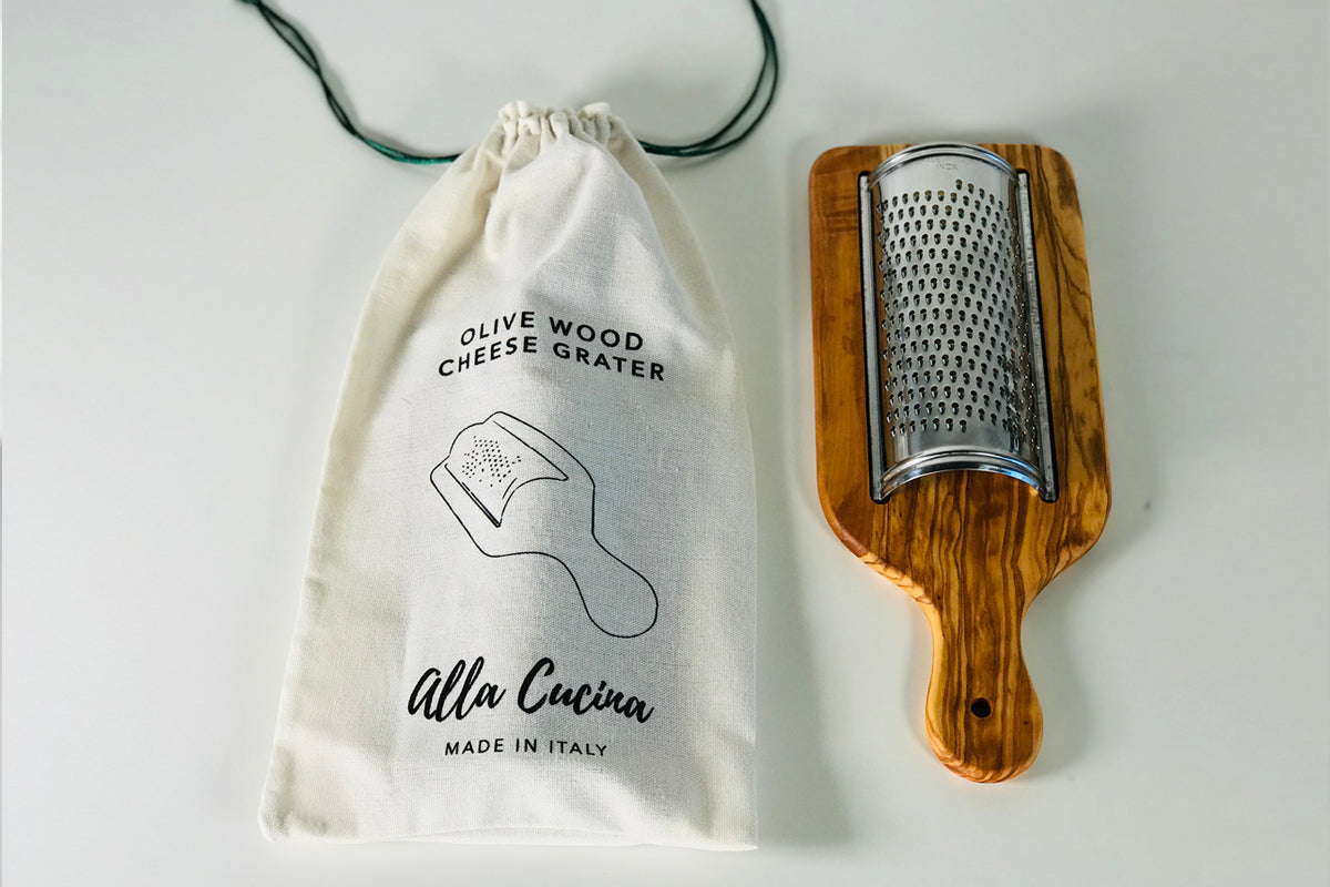 Flat olivewood cheese grater with stainless steel cylindrical grating surface and cotton drawstring storage pouch.
