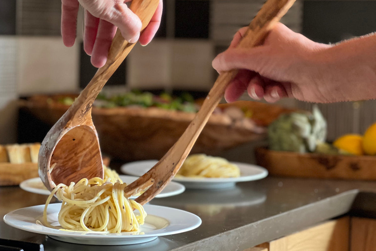 Wooden ladle with long handle being used to serve pasta onto a white plate in a kitchen setting.
