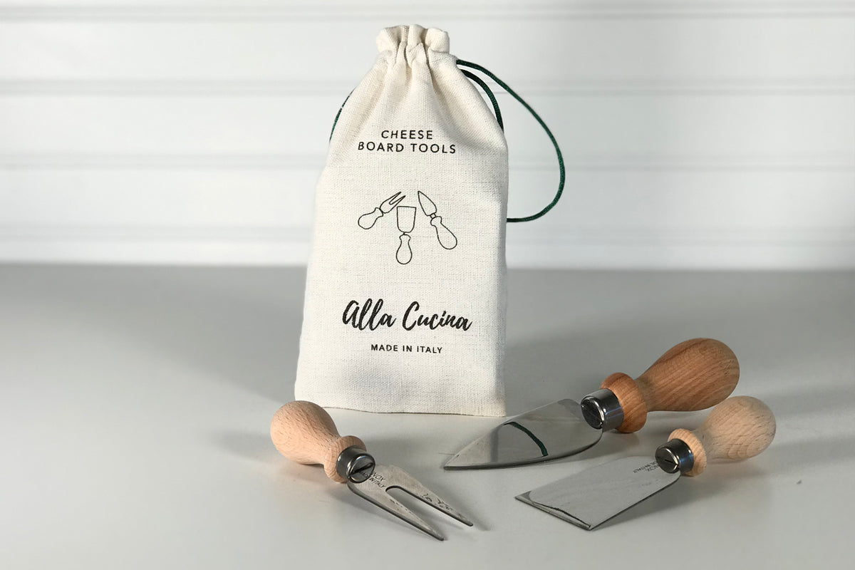 Three cheese knives with wooden handles and metal blades displayed next to a cream linen drawstring bag labeled "Cheese Board