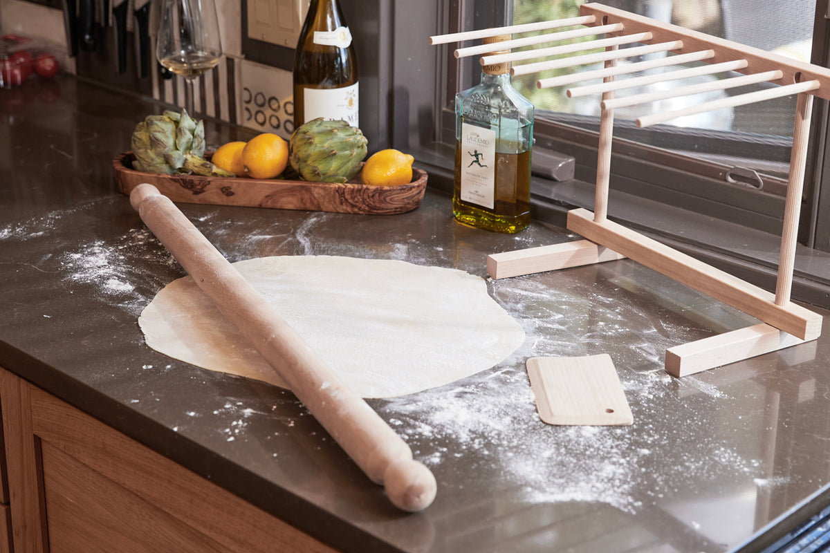 Wooden pasta rolling pin and dough scraper on floured surface with pasta drying rack and kitchen items.
