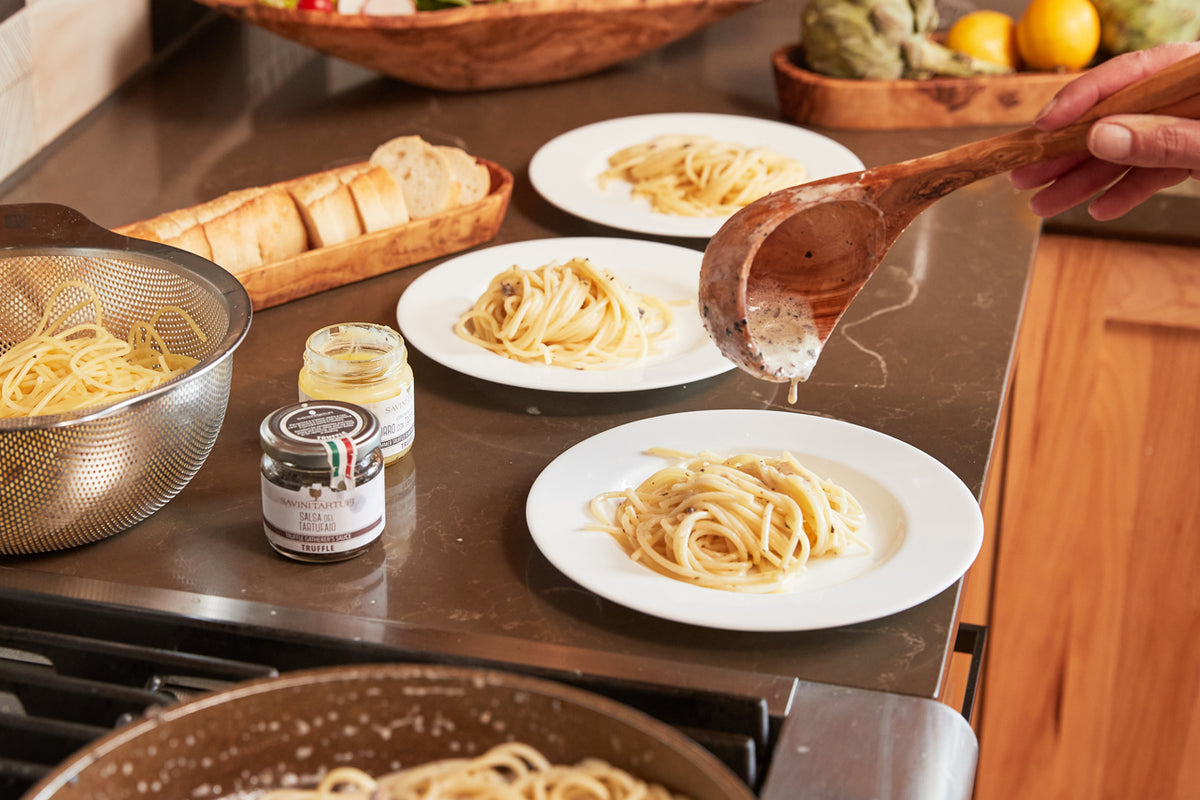 Jar of truffle sauce displayed on kitchen counter with pasta dishes, bread, and cooking equipment.