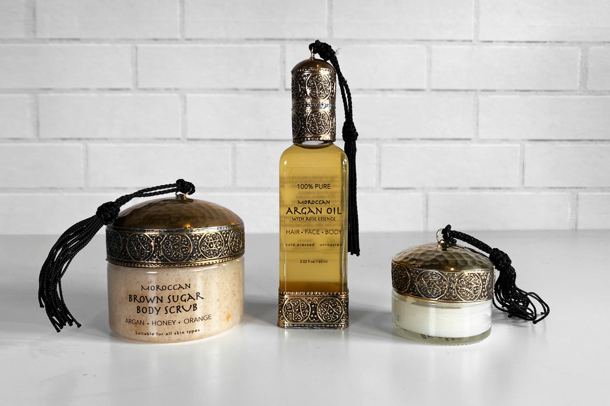 Three Moroccan beauty products with ornate metal lids and black cords: large brown sugar body scrub jar, argan oil bottle, an