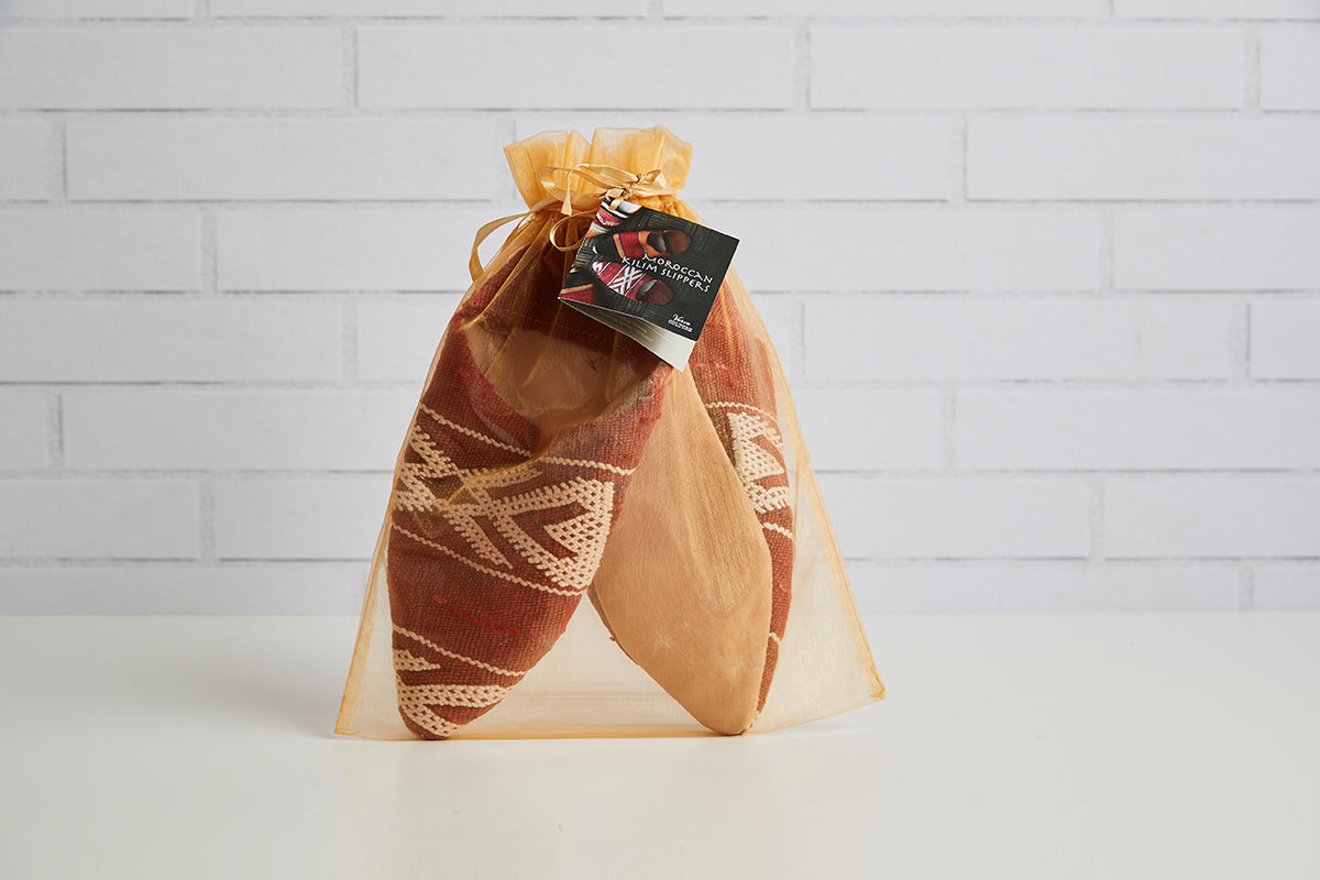 Pair of Moroccan kilim slippers with geometric patterns in rust and cream, enclosed in organza drawstring bag with label tag.