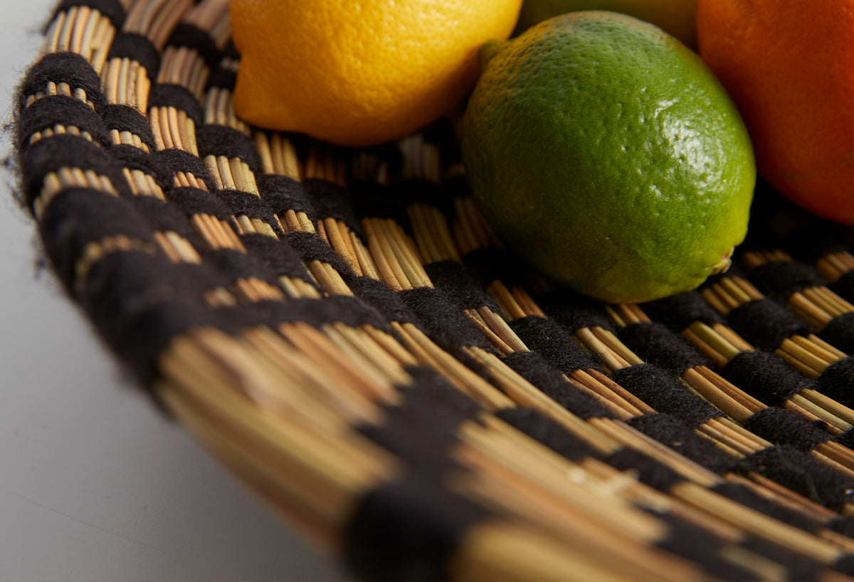 Woven tray with natural straw and black geometric trim holding a green apple, yellow lemon, and orange.
