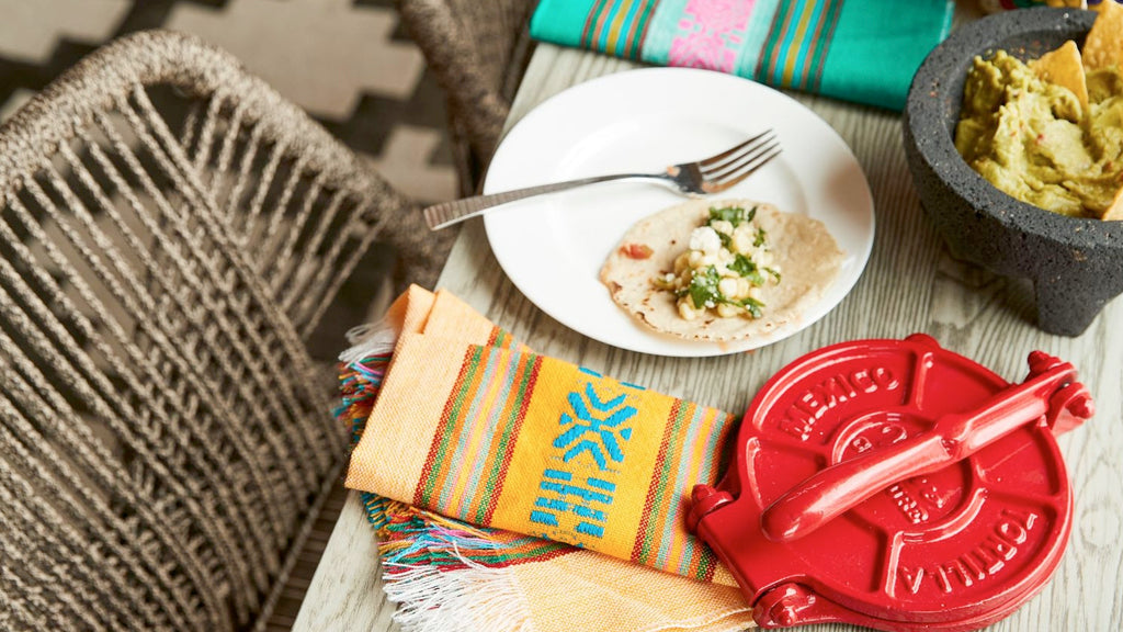 A rustic dining setup featuring a red tortilla press, a freshly made taco on a white plate with a fork, colorful woven napkins, and a traditional stone molcajete filled with guacamole, all set on a wooden table with woven chairs.