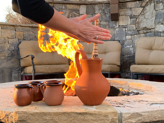 Hand warming over a fire pit beside traditional clay pitchers and mugs outdoors, with a stone wall and patio seating in the background