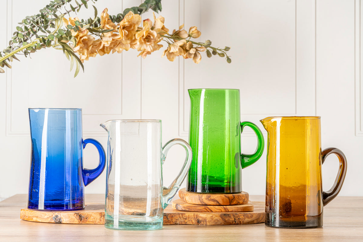 Four cone-shaped glass jugs in blue, clear, green, and amber with handles, displayed on wooden bases.