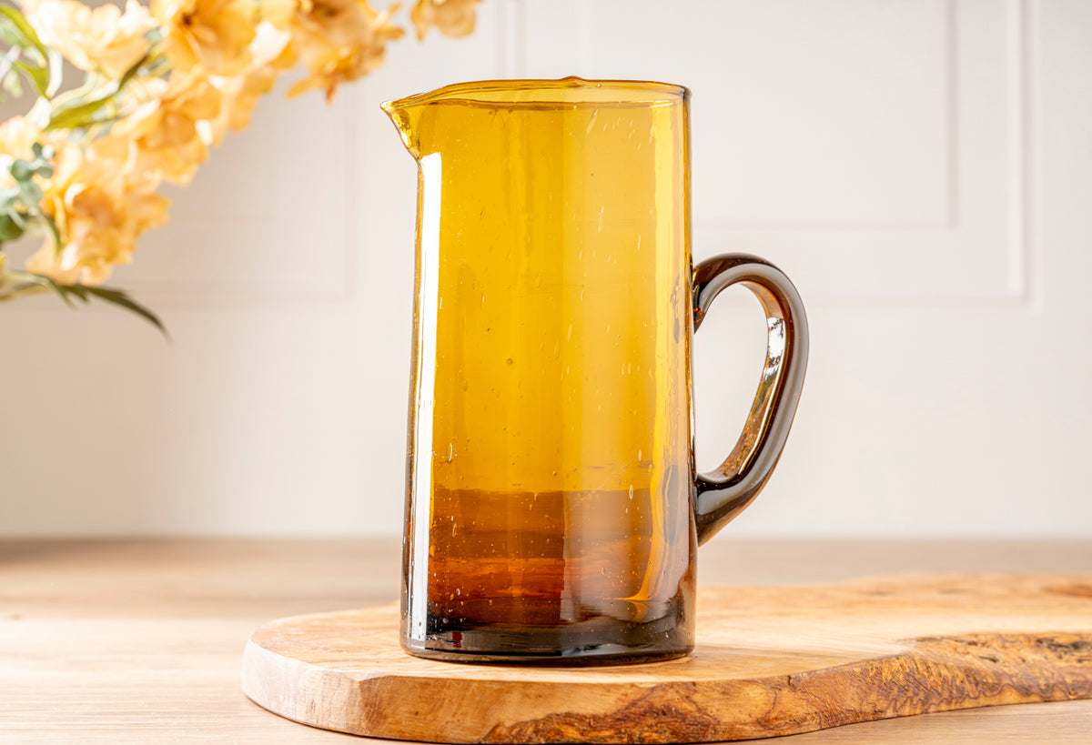 Amber glass jug with cone-shaped body, brown handle, and pouring spout on wooden base.
