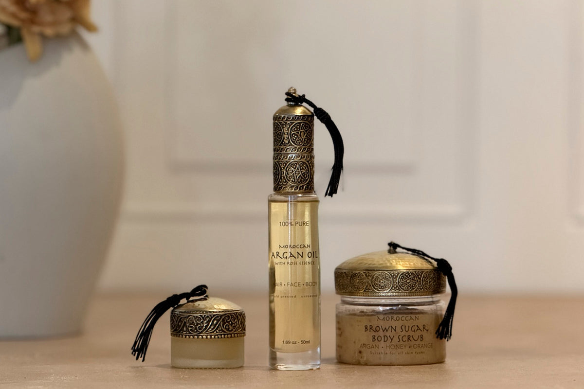Three products with ornate brass caps and black tassel cords: small jar, tall bottle of argan oil, and brown body scrub jar.
