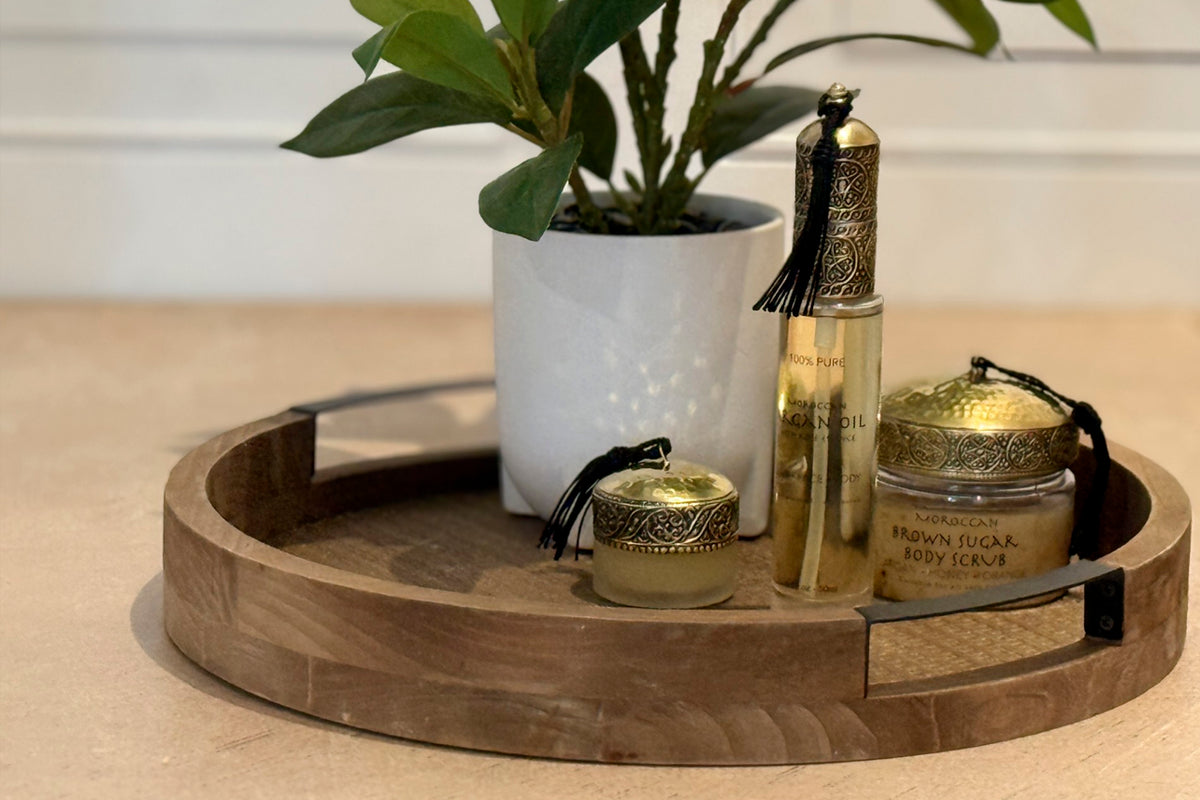 Wooden tray holding argan oil bottle, body scrub jar, small container, and potted green plant with white ceramic pot.