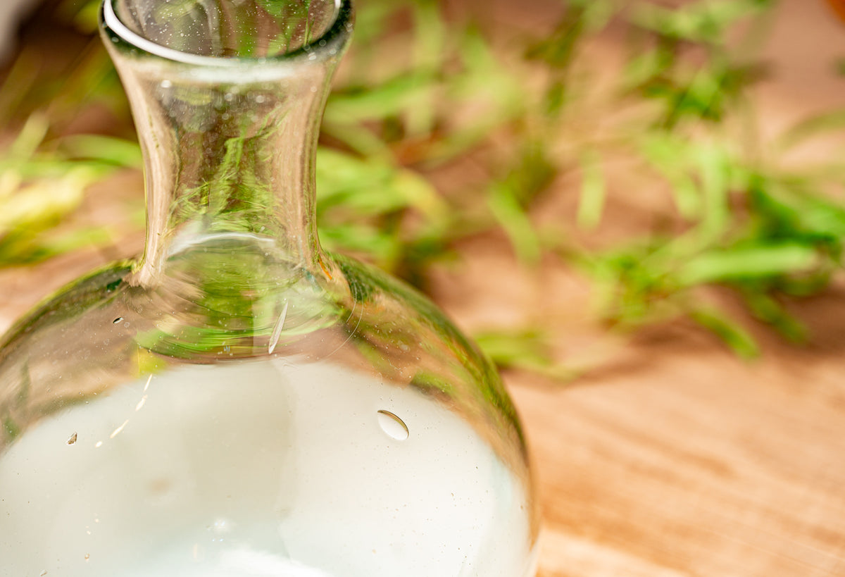 Clear glass carafe with narrow neck and round body containing white liquid, surrounded by green foliage.