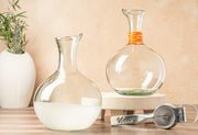 Two clear glass carafes with pour spouts, one containing white liquid, displayed with an orange-wrapped neck detail on the la