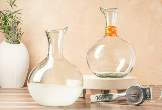 Two clear glass carafes with pour spouts, one containing white liquid, displayed with an orange-wrapped neck detail on the la