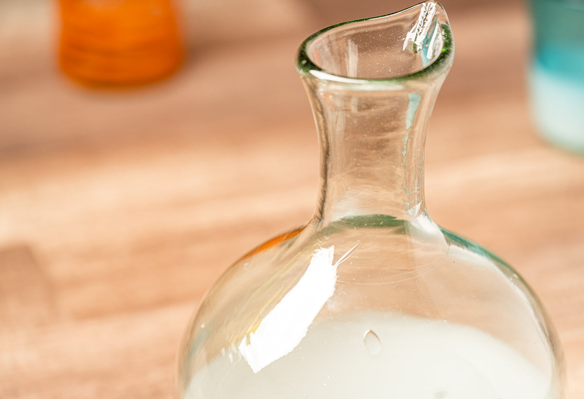 Clear glass carafe with green rim and narrow neck spout, containing white liquid on wooden surface.