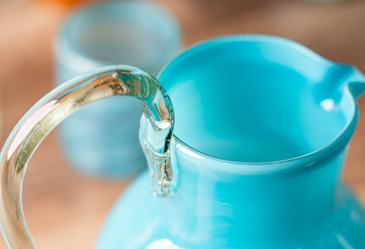 Turquoise glass pitcher with clear glass handle, shown in close-up detail against blurred background.