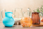 Four glass pitchers in varying sizes and colors: turquoise, clear, orange with textured wrap, and amber.