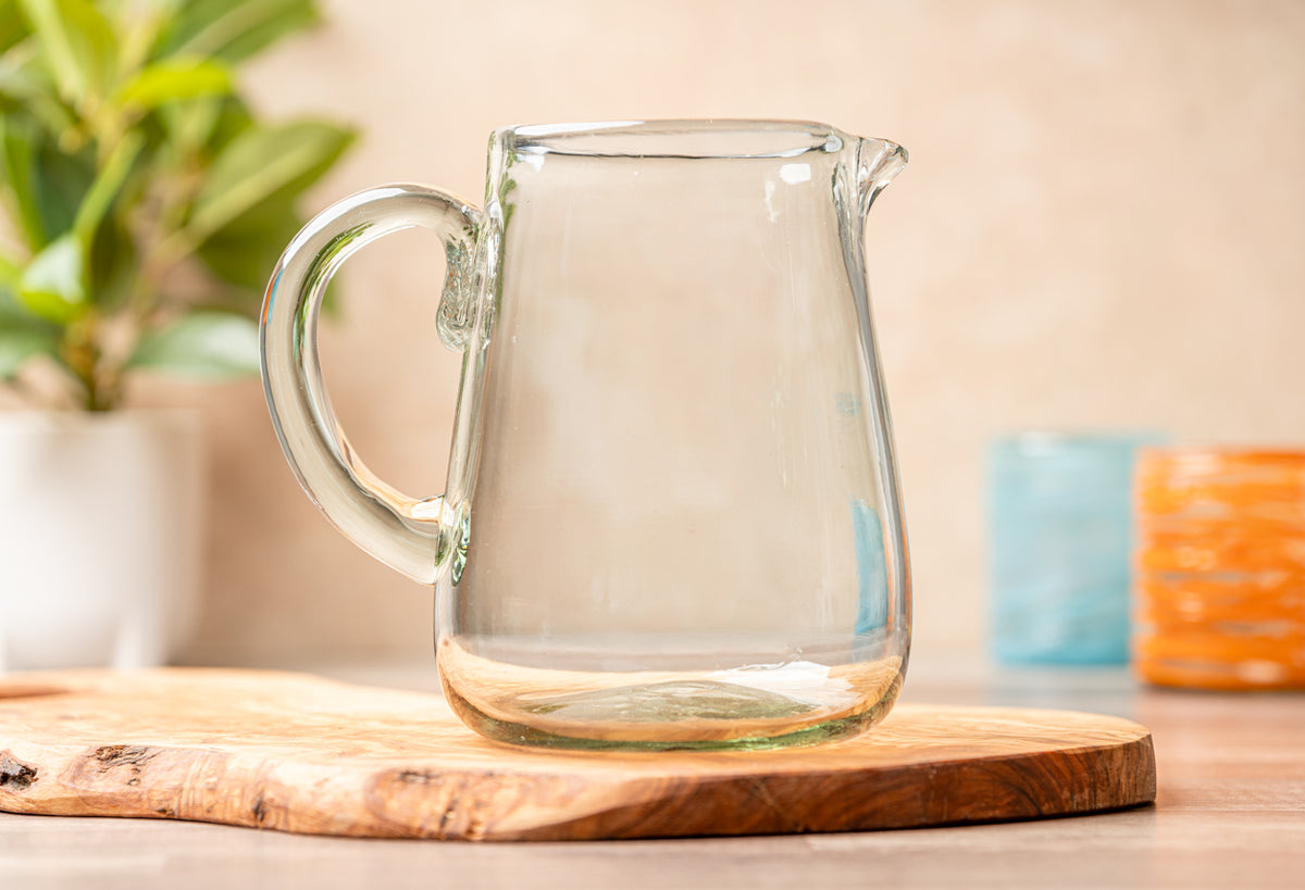 Clear glass pitcher with handle on wooden surface, potted plant and colored objects visible in background.