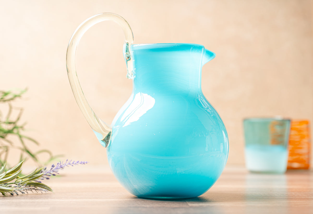 Turquoise glass pitcher with clear handle and pouring spout, displayed on wooden surface.