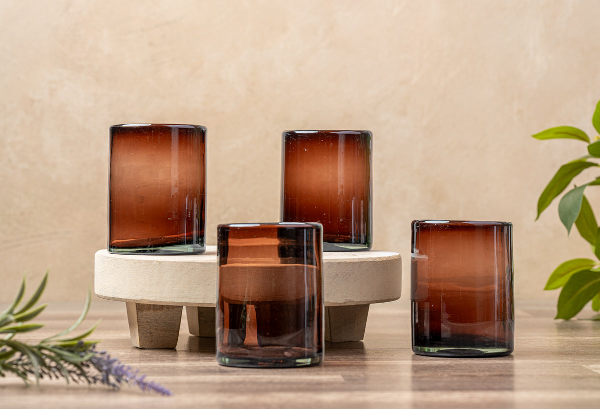 Four tamarind-colored handblown glasses with varying heights displayed on a wooden surface with a concrete holder.