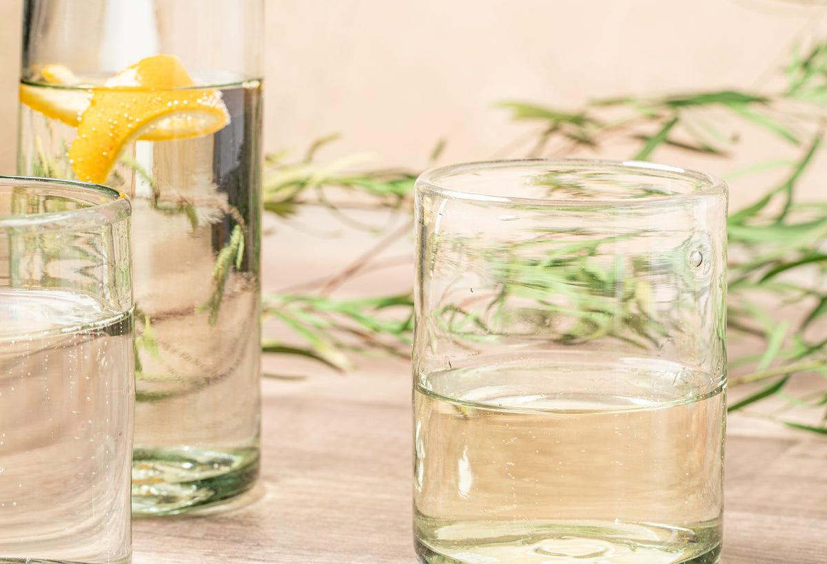 Three clear handblown glasses filled with water and lemon, one containing a lemon slice, on a wooden surface with greenery.